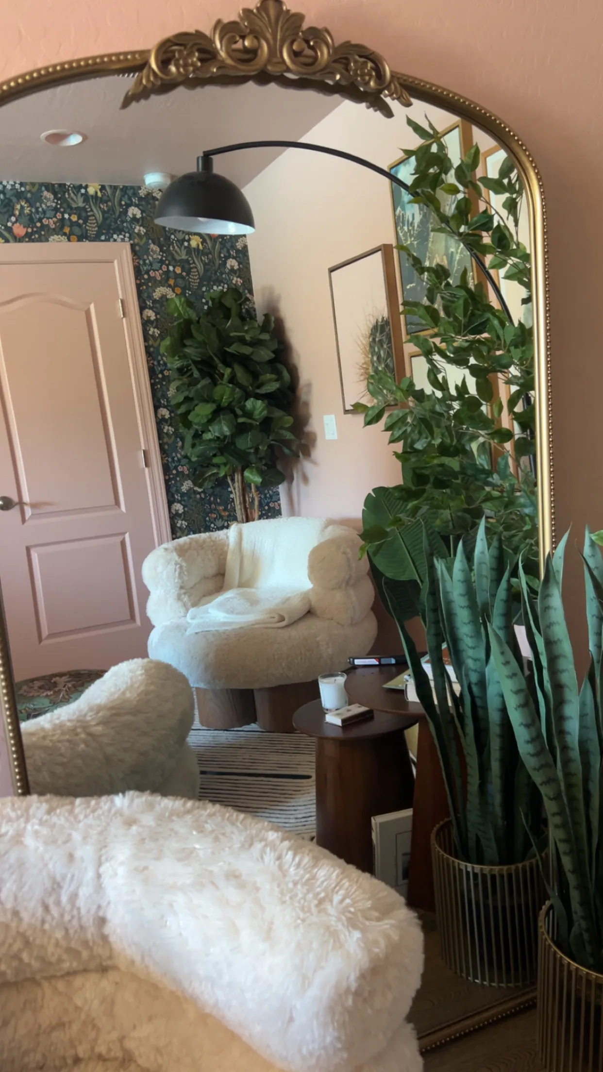 A pink sitting room with green botanical wallpaper seen in a tall arched mirror